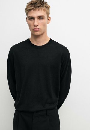 Junger Mann mit kurzen blonden Haaren, der einen schlichten schwarzen Langarmpullover und schwarze Hose trägt und vor einem weißen Hintergrund steht.