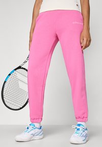 Pink, fleece jogger pants with elastic cuffs and a drawstring waist, featuring a white logo on the thigh. Worn with white and blue tennis shoes.