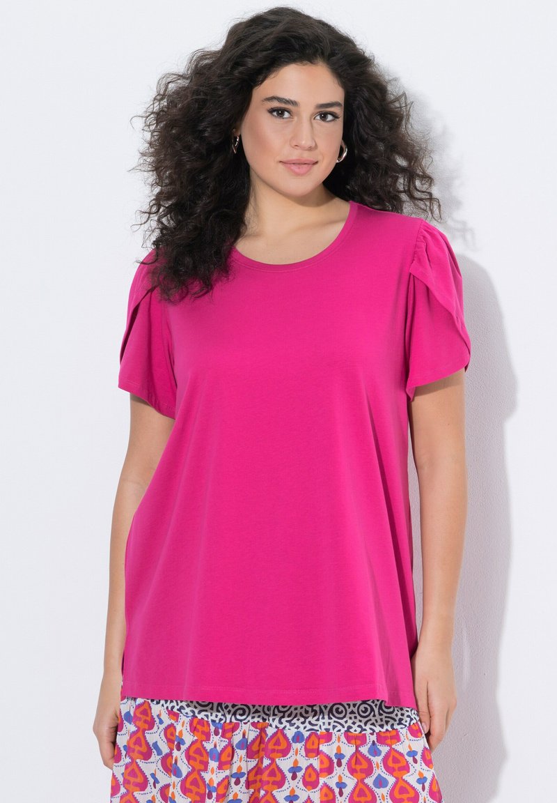 Frau mit lockigem Haar trägt ein knallpinkes Kurzarmoberteil und einen mehrfarbig gemusterten Rock und steht vor einem weißen Hintergrund.