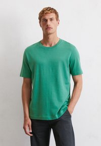 T-shirt vert à manches courtes en tissu doux avec un col rond et une coupe décontractée, associé à un pantalon sombre. Texture lisse.