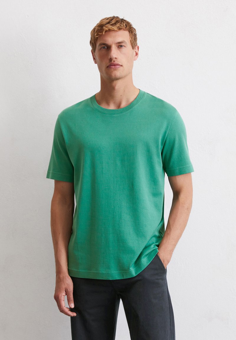 T-shirt vert à manches courtes en tissu doux avec un col rond et une coupe décontractée, associé à un pantalon sombre. Texture lisse.