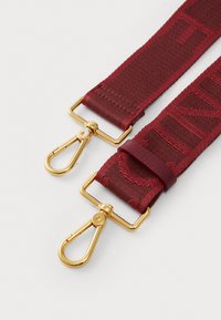 Red textile strap featuring gold metal clips and hardware, with woven lettering detail along the length and a smooth leather accent.