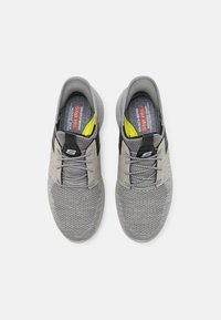 Baskets grises à enfiler avec une tige en mesh, des bandes accent noires, et une semelle texturée. Elles comportent des lacets gris et une doublure intérieure jaune.