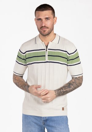 Man met kort haar en tatoeages draagt een witte gebreide poloshirt met groene en zwarte strepen, staand met de handen voor zich gevouwen.