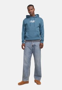Teal hoodie with a front pocket, white logo, and drawstring. Paired with loose-fitting light blue jeans and brown footwear.