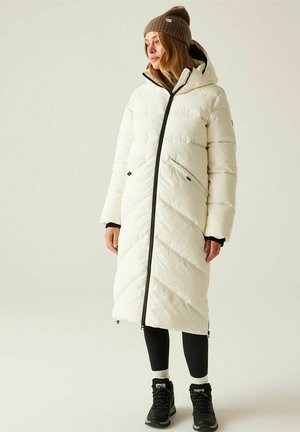 Kobieta ubrana w długi biały pikowany płaszcz z kapturem, brązową dzianinową czapkę, czarne legginsy i czarne buty trekkingowe, stojąca na jednolitym tle.