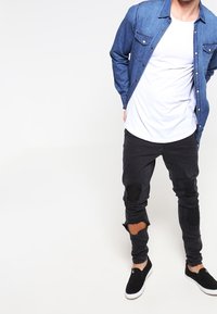 Jasnoniebieska koszula dżinsowa na guziki z kieszeniami; biała koszulka; czarne podarte dżinsy; czarne wsuwane buty. Swobodny strój z wyraźnie widocznymi warstwami.