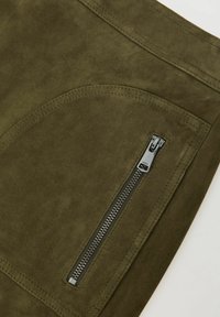 Pantaloni din piele întoarsă verde măsliniu, cu buzunar cu fermoar, cusături plate și textură netedă. Fermoarul are un finisaj metalic argintiu.