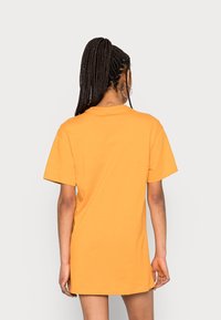 Kvinna med långt flätat hår som bär en oversized orange t-shirt med korta ärmar, som står vänd bort mot en enfärgad ljus bakgrund.
