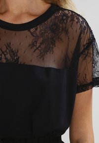 Svart blus med ett genomskinligt spetsoberstykke med blommönster och korta ärmar, i kontrast till en enhetlig svart nedre del.