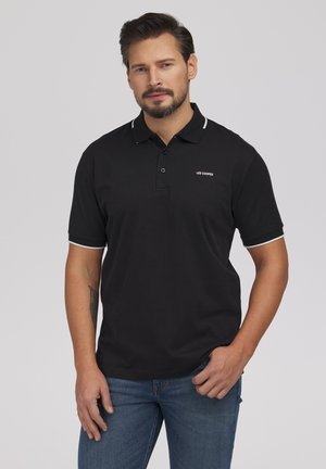 Mężczyzna z brodą w czarnej koszulce polo Lee Cooper z białym obszyciem, stojący na jednolitym szarym tle.