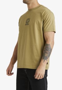 Camiseta de algodón verde oliva con mangas cortas, escote redondo y un diseño gráfico central. Presenta un corte recto y pequeñas aberturas laterales.