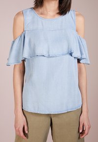 Ljusblå kallaxel blus med volanger, bärs med olivgröna byxor, visad från halsen till höfterna mot en neutral bakgrund.