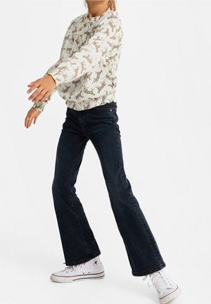 Mujer con blusa blanca de estampado de leopardo, jeans oscuros acampanados y zapatillas Converse blancas, posando con los brazos extendidos hacia adelante.