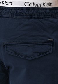 Shorts bleu marine en tissu texturé, avec une poche arrière et une ceinture élastique blanche portant l'étiquette "Calvin Klein".