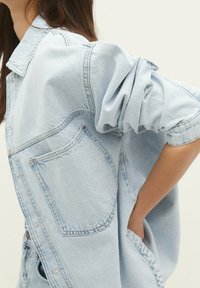 Helles blaues Jeanshemd mit klassischem Kragen, großen Fronttaschen und hochgekrempelten Ärmeln, mit lässiger Passform und leicht strukturiertem Material.