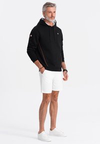 Sudadera negra con costuras naranjas, pantalones cortos blancos y zapatillas blancas. La sudadera tiene un cordón y un logo en el pecho.