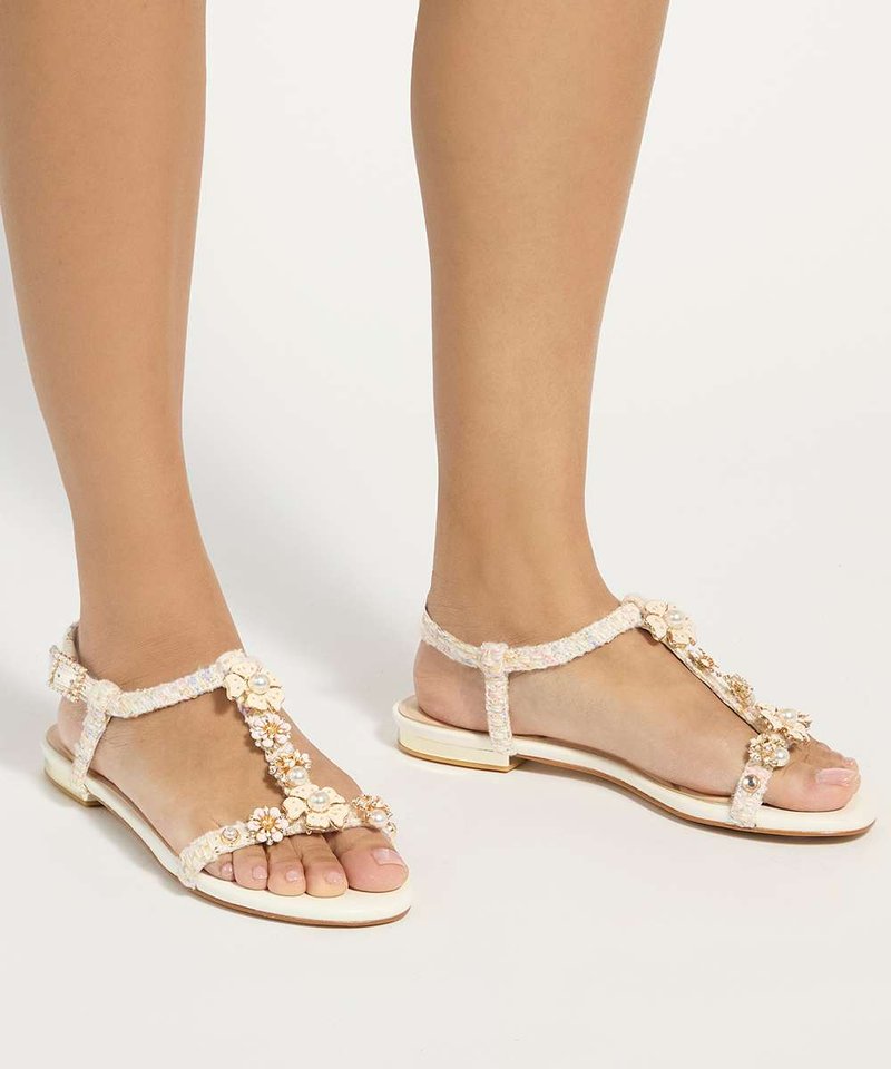 Pieds portant des sandales plates blanches avec des brides tressées et des fleurs décoratives ainsi que des perles sur un fond neutre.