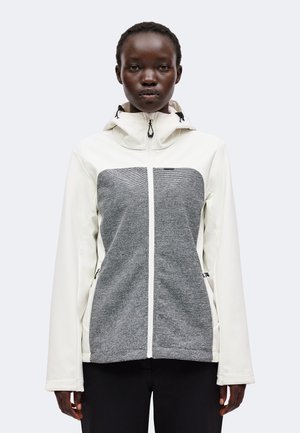 Femme portant une veste blanche à capuche avec un panneau gris texturé à l'avant, debout, droite, devant un fond clair uni.