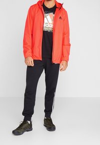 Red lightweight jacket with hood, black graphic t-shirt, black sweatpants with white stripes, and dark green athletic shoes.