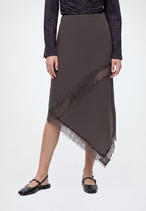 Personne portant une jupe asymétrique sombre avec bordure en dentelle, une chemise à manches longues à carreaux sombres et des chaussures Mary Jane noires.