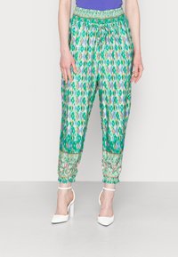 Calças jogger verdes com um padrão, cintura com cordão e punhos elásticos. Apresenta uma faixa floral decorativa na bainha. Combinadas com sapatos de salto brancos.