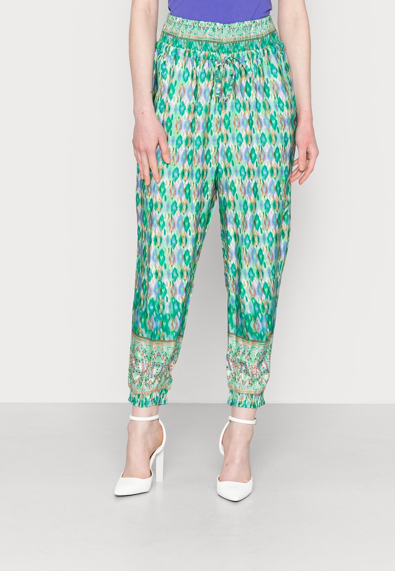 Calças jogger verdes com um padrão, cintura com cordão e punhos elásticos. Apresenta uma faixa floral decorativa na bainha. Combinadas com sapatos de salto brancos.