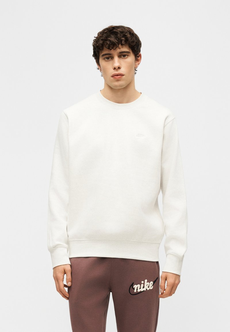 Giovane uomo con capelli ricci scuri che indossa una felpa bianca semplice e pantaloni della tuta marroni Nike con logo sulla coscia sinistra.