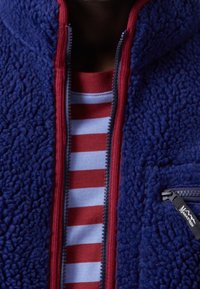 Blå fleecejakke med røde detaljer og en lynlåslomme, over en stribet rød og blå t-shirt. Blød tekstur, høj krave og lynlåsdetalje.