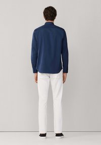 Granatowa koszula z długim rękawem i kołnierzykiem, zestawiona z białymi spodniami i ciemnymi butami. Gładki materiał o luźnym kroju; widoczne detale z guzikami na mankietach.