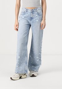 Jeans de pierna ancha en azul claro con detalles desgastados y dobladillo deshilachado. Fabricados en denim con rotos visibles y un ajuste relajado, combinados con zapatillas.