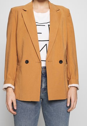 Personne portant un blazer jaune moutarde à double boutonnière sur un t-shirt blanc à motif graphique et un jean bleu, avec les manches du blazer retroussées.