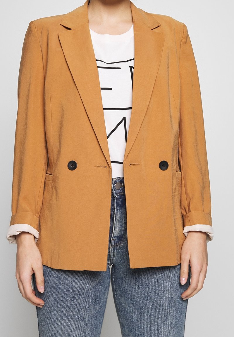 Personne portant un blazer jaune moutarde à double boutonnière sur un t-shirt blanc à motif graphique et un jean bleu, avec les manches du blazer retroussées.
