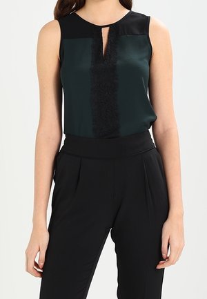 Femme portant un chemisier sans manches vert foncé avec bordure en dentelle noire et un pantalon plissé noir taille haute, debout, bras détendus.