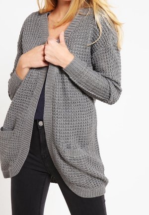 Femme portant un cardigan en maille gris texturé par-dessus un haut sombre et un pantalon noir, tenant le cardigan fermé au niveau de la poitrine.