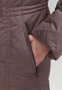 Brun vadderad jacka med sidoficka med dragkedja, elastisk midja och slät, glänsande tygtextur. Accenter inkluderar sömnadsdetaljer.