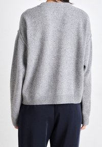 Maglione grigio a maglia con vestibilità rilassata, orlo e polsini a costine, che mostra una finitura morbida e texture. Indossato sopra pantaloni scuri a gamba dritta.