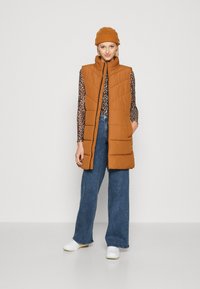 Brun puffer vest med høy krage, frontlås og skrå quilting. Kombinert med et mønstrete topp og vide jeans.