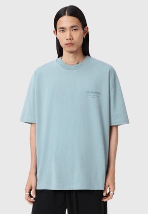 Młoda osoba z długimi czarnymi włosami nosi oversize jasnoniebieski T-shirt AllSaints i czarne spodnie, stoi na jednolitym tle.