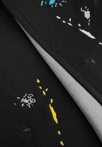 Zwart patchwork stof toont meerkleurige geborduurde patronen in blauw, geel en zilver, met een gladde grijze voering zichtbaar.