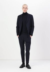 Navy blazer with matching pants, worn over a black turtleneck. Fabric appears smooth; outfit features tailored fit and minimalistic design.