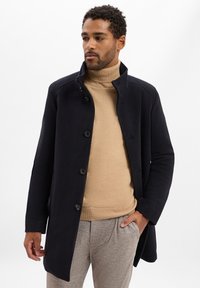 Marineblauer Wollmischmantel mit hohem Kragen, fünf Knöpfen und seitlichen Taschen, getragen über einem beigen Rollkragenpullover und hell gemusterten Hosen.