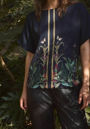 Vrouw in een marineblauwe blouse met verticale gouden strepen en een botanische print met vlinders, bladeren en bloemen, gecombineerd met zwarte broek.