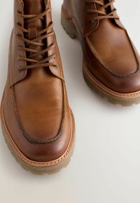 Brązowe skórzane buty z okrągłym noskiem, z przeszyciem i sznurowanym zapięciem, spoczywające na jasnej, teksturowanej powierzchni. Widoczne są gumowe podeszwy.