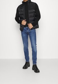 Chaleco de plumas negro con cuello alto, bolsillos frontales con cremallera, combinado con un suéter de canalé negro, emparejado con jeans ajustados azules y botas negras.