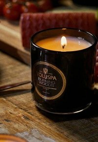 Svart glasljushållare med guldetikett som visar "VOLUSPA BURNING WOODS." Innehåller gul vax, tänd veke, placerad på en träyta.