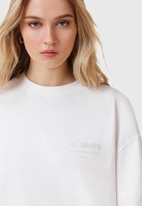 Biała bluza z okrągłym dekoltem, krótkimi rękawami i subtelnym, refleksyjnym logo "ALLSAINTS" na piersi. Miękka faktura materiału.