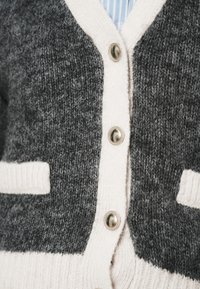 Gray and cream cardigan with a textured knit, featuring a v-neck, three metallic buttons, and two cream pockets.