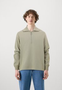 Lys grøn glidelåsgenser med høyt krage, ribbede mansjetter og en glatt tekstur; kombinert med blå denimjeans.
