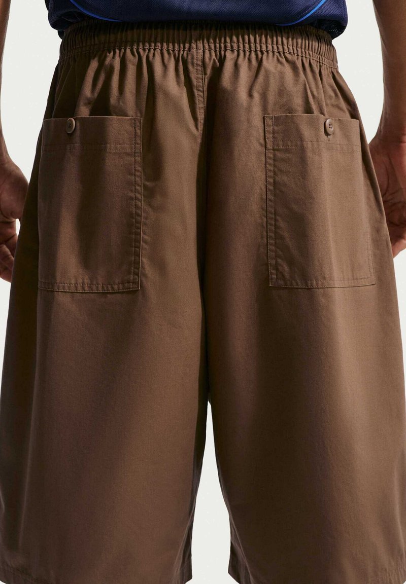 Pantalones cortos marrones de corte ancho con dos bolsillos traseros abotonados, usados por una persona de pie frente a un fondo liso.